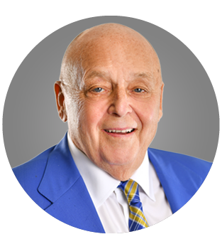 Portrait of an elderly man wearing a blue blazer, white shirt, and yellow plaid tie, smiling against a gray background.