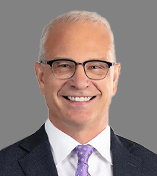 Portrait of a smiling man with glasses and gray hair, dressed in a dark suit, white shirt, and purple tie, against a gray background.
