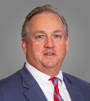 A middle-aged man with light skin, gray hair, and blue eyes, dressed in a blue suit, white shirt, and red striped tie, posing against a plain gray background.